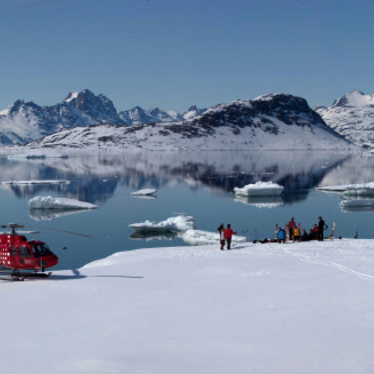 Heli skiers taking a lunch break next to a mirror like fjord on the east coast of Greenland with the surrounding mountains mirrored in the calm waters.
