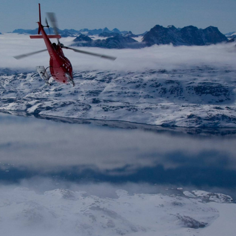 Heli-skiing helicopter flying towards a fjord on the east coast of Greenland. Majestic snow capped mountains rising out of the mist in the distance.