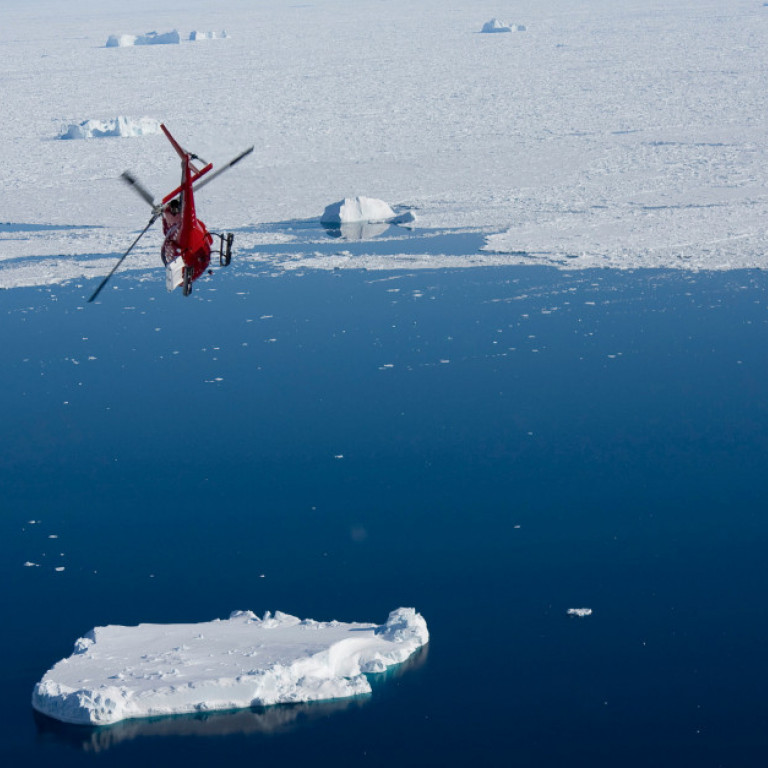 Heli-skiing helicopter descending towards the open waters on Greenland’s east coast to pick up another group of heli-skiers with the pack ice in the background.
