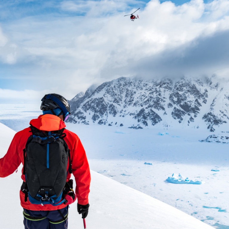 East Greenland heliskiing. Skier gazing over a frozen ice filled fjord whilst a heli-skiing helicopter approaches. Icebergs and pack ice as far as one can see.  ©Eli Spiegel